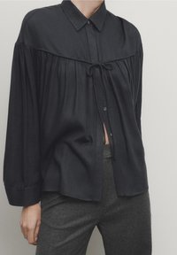 Zwarte button-up shirt met kraag, gebundelde voorkant met een strikdetail en lange mouwen; zachte textuur en relaxed fit.