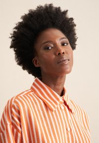 Camicia a righe arancioni e bianche con colletto e chiusura a bottone, realizzata in un tessuto leggero; presenta una vestibilità rilassata e una texture liscia.