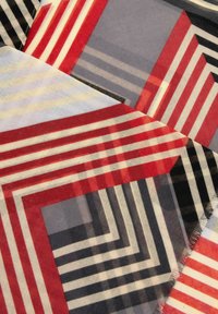 Tessuto geometrico con strisce sovrapposte in rosso, nero, grigio e beige, dalla texture morbida e bordi sfilacciati.