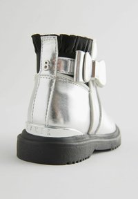 Metallisch silberner Knöchelstiefel mit einem schwarzen elastischen Kragen, großer Schleifenakzent und einer strukturierten schwarzen Sohle. Branding im Absatz eingeprägt.