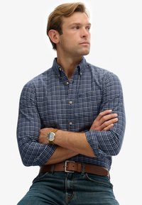 Hombre con los brazos cruzados, vistiendo una camisa de cuadros azul, reloj de cuero marrón, cinturón marrón y jeans azules, mirando hacia un lado.
