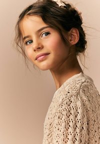 Cardigan a maglia in beige chiaro con motivo a disegno aperto e accenti di paillettes. Tessuto morbido, scollatura rotonda e capelli acconciati in modo sciolto.