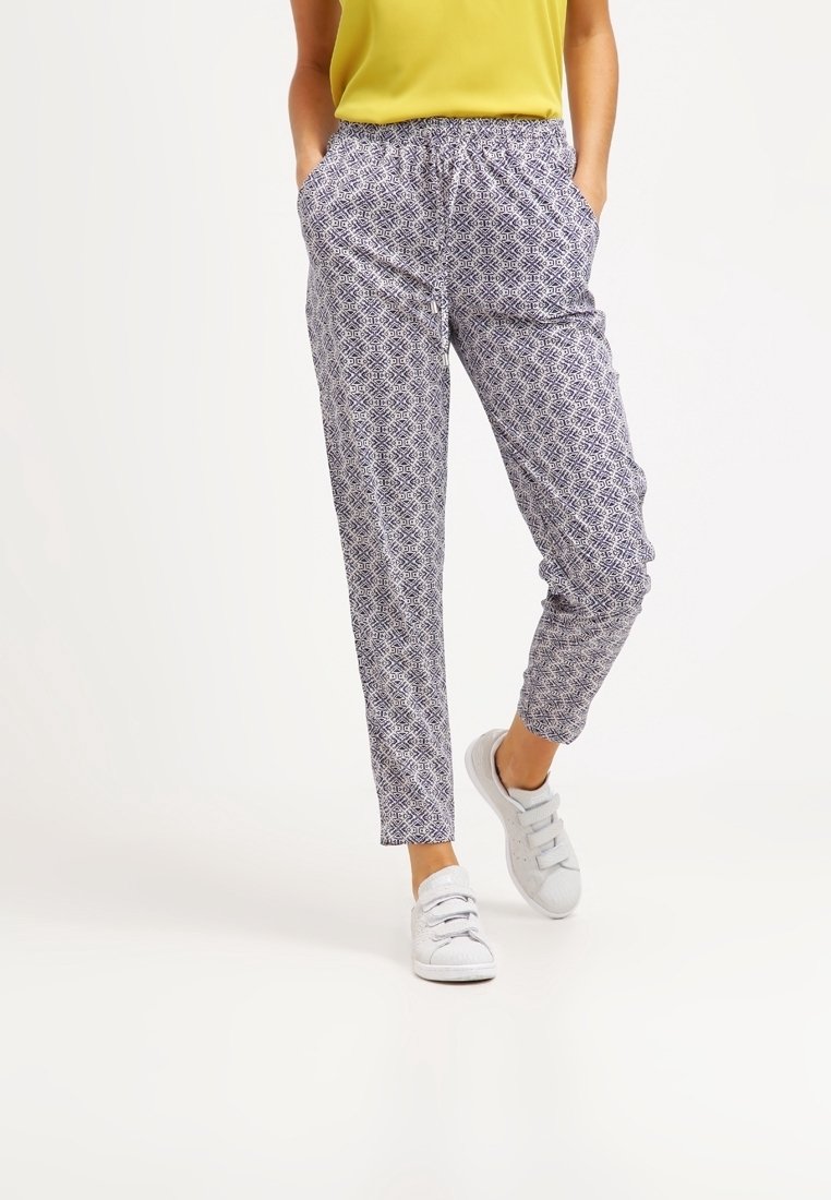 Personne portant un pantalon slim à motifs, un haut jaune et des baskets blanches à scratch, debout les mains dans les poches sur un fond uni.
