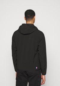 Mann in einer schwarzen Kapuzenjacke und schwarzen Cargo-Hosen, mit dem Rücken zu einem hellgrauen Hintergrund.