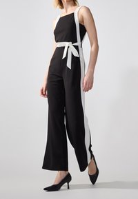 Černý overal s bílými bočními panely, čtvercovým výstřihem a uvázaným páskem. Jednoduchý, elegantní design s širokými nohavicemi a hladkým povrchem.