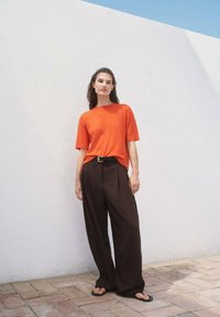 Oranges T-Shirt mit kurzen Ärmeln, kombiniert mit hochtaillierten, locker sitzenden, dunkelbraunen Hosen, mit einem schwarzen Gürtel und flachen Sandalen. Weißer Wandhintergrund.