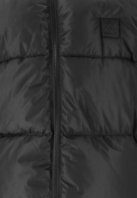 Doudoune noire avec fermeture éclair, design matelassé, texture lisse et un petit logo brodé sur la poitrine.
