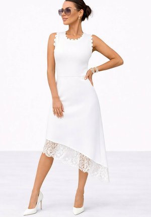 Femme portant une robe blanche sans manches avec des bords festonnés et un ourlet en dentelle asymétrique, assortie à des talons blancs et des lunettes de soleil.