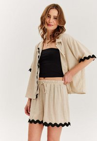 Svetle béžový lněný set s krátkým rukávem, obsahující košili na knoflíky s černým zvlněným lemem a kraťasy s elastickým pasem, doplněný černým topem bez ramínek.