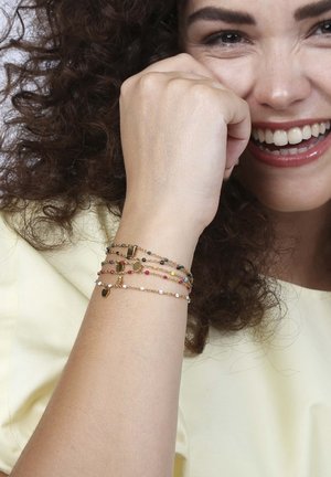 Gouden armbanden met kleurrijke kralen in rood, zwart en wit, gestapeld op een pols tegen een lichtgele achtergrond. Gladde textuur.
