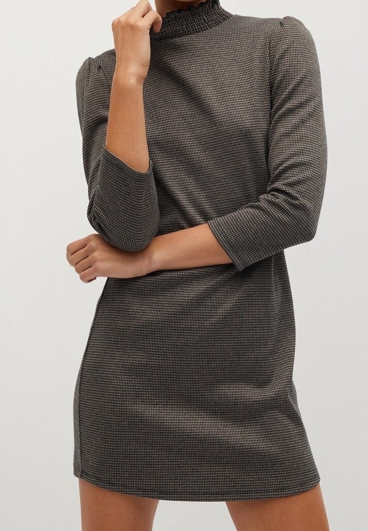 Femme portant une mini-robe ajustée à manches longues à carreaux noirs et blancs avec un col montant à volants, posant les bras croisés.