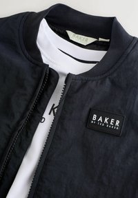 Juodas švarko tipo užtrauktukas su Baker by Ted Baker logotipo pleistru, dėvimas virš baltos palaidinės su dalinai matomu juodu tekstu.