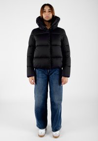 Giacca a puno nera con colletto alto e cappuccio, caratterizzata da sezioni trapuntate, abbinata a jeans blu a vestibilità ampia e sneakers bianche.