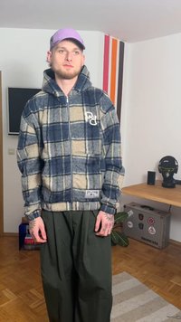 Jakna iz flisa z zadrgo, z mornarsko modrim in bež karo vzorcem, s kapuco in logotipom. Kombinirana z ohlapnimi olivno zelenimi hlačami, ki razkrivajo zapletene tetovaže.