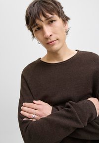 Ung person med kort brunt hår iført en mørkebrun tekstureret sweater, sølv hoopsmykker og ringe, der krydser armene foran en ensfarvet baggrund.