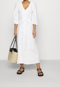 Abito midi bianco con maniche a sbuffo, ricamo floreale testurizzato, scollo a V profondo e vita con laccetto. Indossato con una borsa nera e sandali.