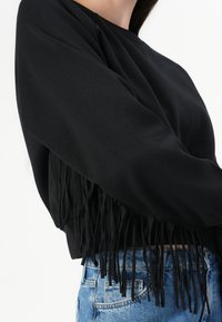 Felpa nera corta con dettagli a frange lungo l'orlo, realizzata in tessuto morbido. Abbinata a pantaloncini di jeans blu chiaro.