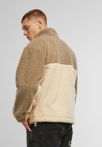 Jachetă din fleece în nuanțe de bej și crem, cu guler înalt, închidere cu fermoar, buzunare laterale și un material moale, texturat pentru căldură.