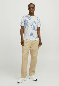 Grå t-shirt med kort ärm och blå lövmönster, tillsammans med beige byxor och vita sneakers. Modellens bär mörka solglasögon.