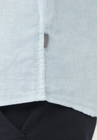 Camisa de tela azul claro con textura suave, que presenta una etiqueta en la costura lateral y un dobladillo recto, combinada con pantalones oscuros.