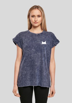 Femme portant un t-shirt bleu marine délavé avec des manches retroussées et un petit motif de chat blanc sur la poitrine gauche, assorti à un pantalon noir.
