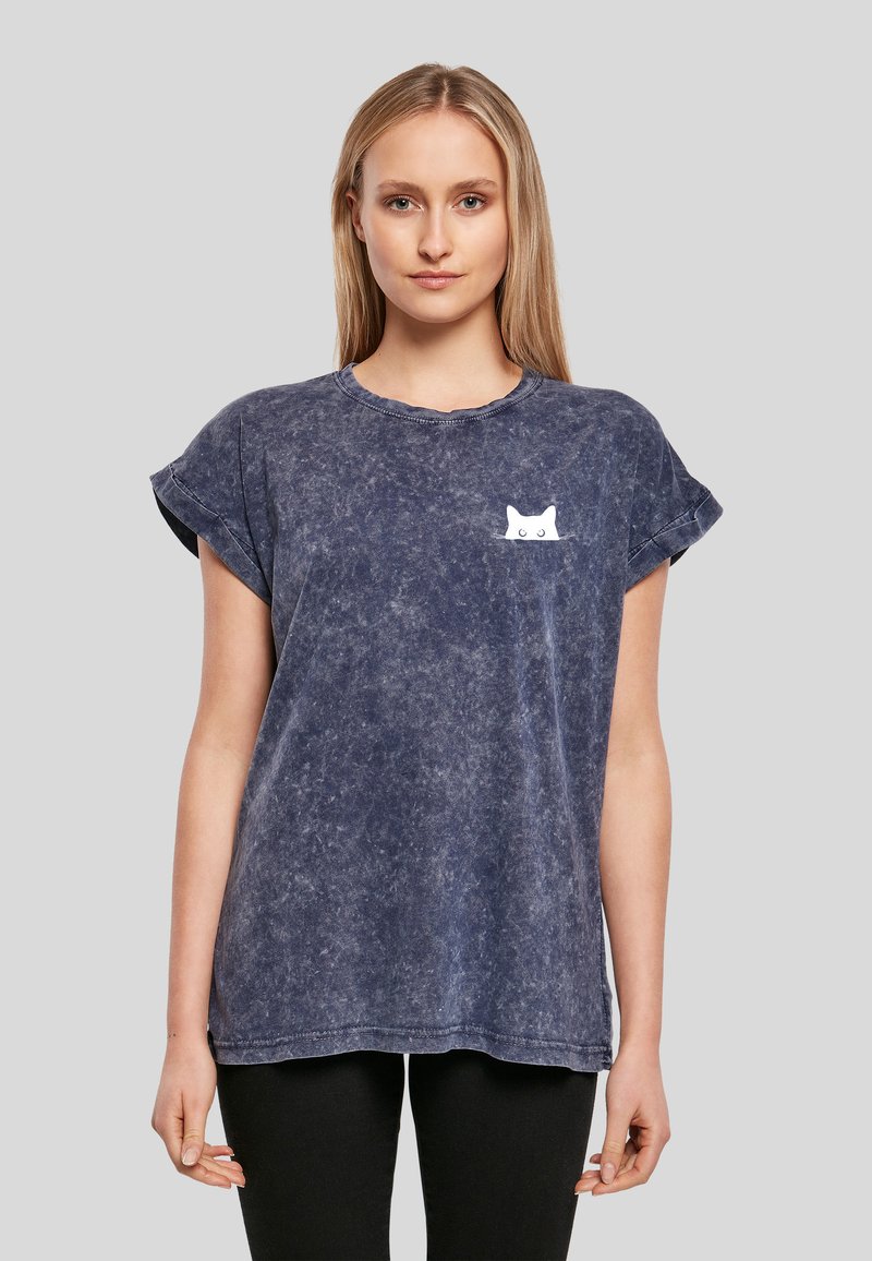 Femme portant un t-shirt bleu marine délavé avec des manches retroussées et un petit motif de chat blanc sur la poitrine gauche, assorti à un pantalon noir.