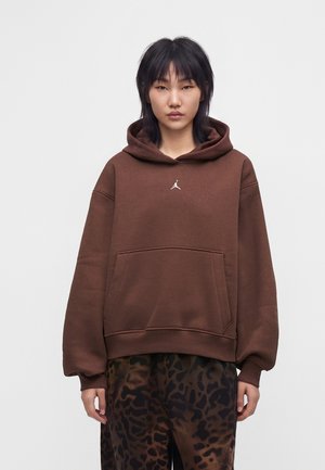 Mujer con sudadera con capucha marrón y pantalones anchos de estampado de leopardo marrón oscuro, de pie con una mano en el bolsillo y expresión neutra.