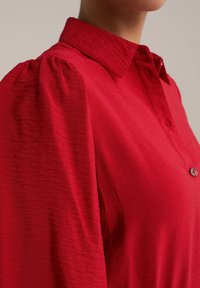 Blouse rouge avec un col boutonné et des manches bouffantes. Le tissu présente un motif texturé, renforçant son attrait visuel et sa profondeur.