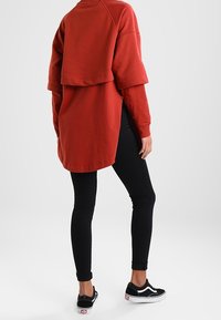 Sudadera roja de gran tamaño con un diseño en capas, con cuello redondo y mangas caídas, combinada con jeans negros ajustados y zapatillas negras.