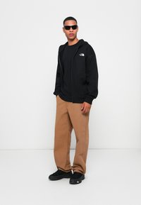 Black zip-up hoodie with logo, black t-shirt, tan wide-leg pants, and black sneakers. Model wearing dark sunglasses, standing against a white backdrop.