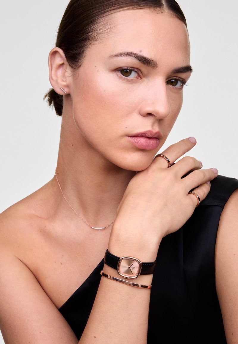 Montre en cuir noir avec un cadran carré en rose doré, accompagnée d'un bracelet fin en rose doré et de bagues, posée sur une épaule nue.
