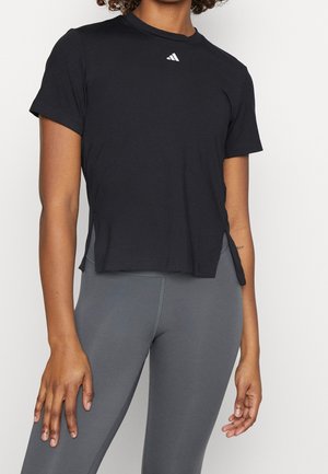 Person trägt ein schwarzes kurzärmeliges Adidas-Sportshirt mit Seitenschlitzen und grauen Leggings mit hohem Bund, steht vor neutralem Hintergrund.