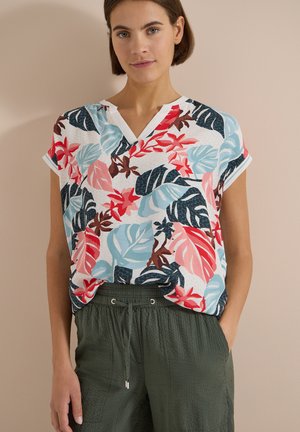 Femme portant un haut blanc à manches courtes avec un imprimé de feuilles tropicales rouges, bleues et brunes, associé à un pantalon vert foncé à cordon.