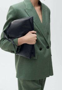 Blazer de lino verde con escote en V profundo, diseño de doble botonadura, combinado con un clutch de cuero negro. Texturas suaves y ajuste a medida.