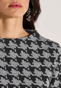 Grijs en zwart hondenstoothpatroon stof met een hoge halslijn, gecombineerd met een opvallende hanger met cirkelvormige en geometrische vormen.