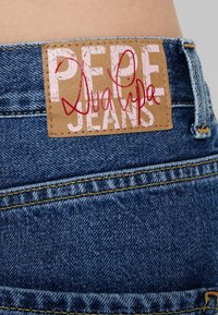 Vaalean sinisessä pesussa olevat farkut, joissa on takana ommeltu ruskea nahkainen merkki, jossa lukee "PEPE JEANS" ja "Dua Lipa" vaaleanpunaisella.