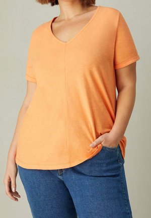Persoon met een oranje T-shirt met V-hals en korte mouwen en een blauwe spijkerbroek, met één hand in de broekzak, bijgesneden van schouders tot heup.
