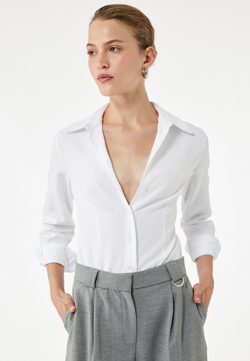 Witte button-up shirt met een V-hals en opgerolde mouwen, gecombineerd met grijze high-waisted broek met riemlus en plooien.