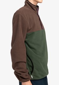 Sudadera de forro polar de capa intermedia con la parte superior marrón, la parte inferior verde y acentos morados. Cuenta con un cuello alto y bolsillos laterales.