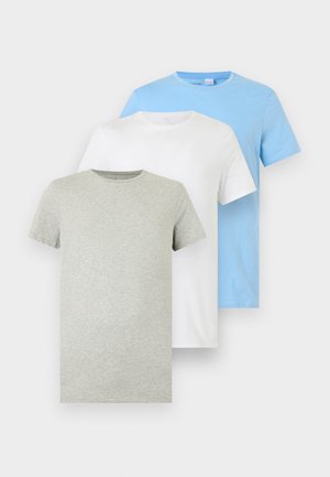 Mees seisab paljajalu, kandes heledat halli lühikeste varrukatega T-särki ja sobivat aluspesu ühevärvilise valge tausta ees.