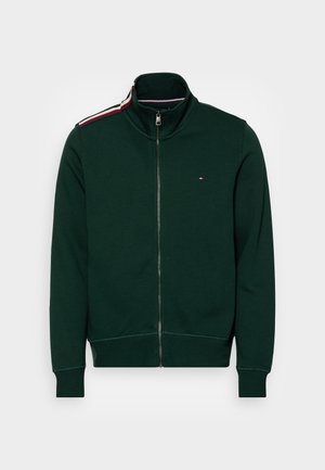 Sweatshirt vert à fermeture éclair avec un col montant, présentant une bande contrastante rouge et blanche sur l'épaule et un petit logo sur la poitrine.