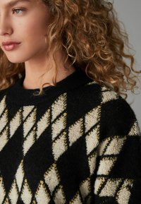 Pull noir avec un motif en losanges crème et jaune, présentant une finition texturée et un col côtelé. Montre un gros plan du col et du design.