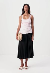 Top in cotone rosa pallido a costine con scollatura ondulata, abbinato a una gonna lunga nera plissettata e sandali a scivolo crema, mentre tiene in mano una borsa beige.