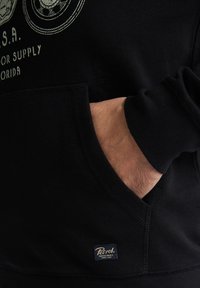 Felpa nera con tasca a marsupio, caratterizzata da un scollo a V e polsini a costine. Include una piccola etichetta del marchio con il logo "Petrol Industries".