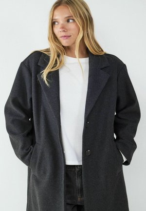 Donkergrijze wollen jas met een klassieke revers, enkelvoudig ontwerp en twee knopen. Bevat zijzakken en heeft een ontspannen pasvorm.