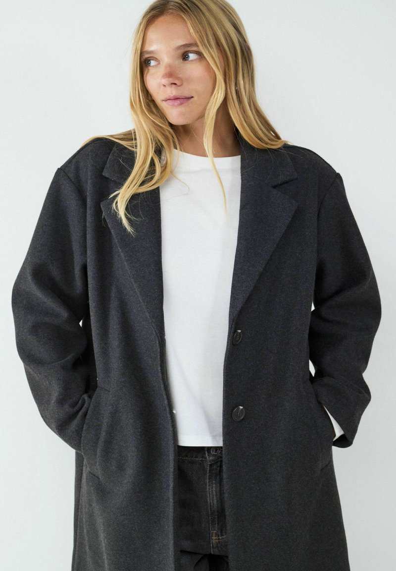 Darker grauer Wollmantel mit klassischem Revers, einreihigem Design und zwei Knöpfen. Verfügt über Seitenflecken und eine lockere Passform.