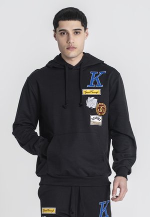Sudadera negra de algodón, con un bolsillo canguro. Parche bordado en azul, amarillo y blanco. Incluye capucha con cordón.