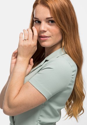 Blusa verde chiaro a maniche corte con una consistenza morbida. Modella che indossa un anello d'argento, mostrando capelli ricci e rossi e un'espressione neutra.