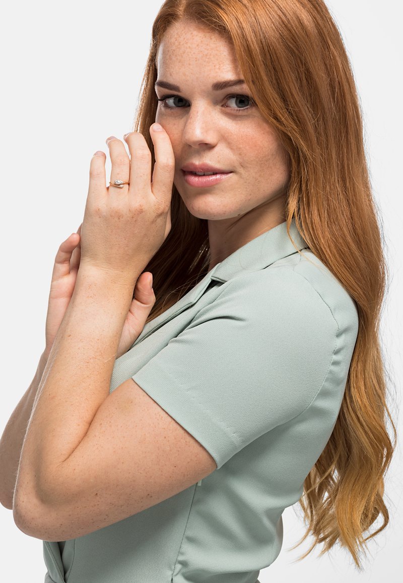 Blusa verde chiaro a maniche corte con una consistenza morbida. Modella che indossa un anello d'argento, mostrando capelli ricci e rossi e un'espressione neutra.