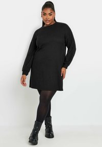 Abito in maglia nera a coste con maniche lunghe e vestibilità comoda, caratterizzato da un collo rotondo e lunghezza a metà coscia, abbinato a stivali neri alla caviglia.